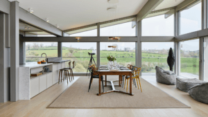 Open living space overlooking field of cows. Various artistic elements including colourful chairs, floor to ceiling windows and a anthropormorphic bird man
