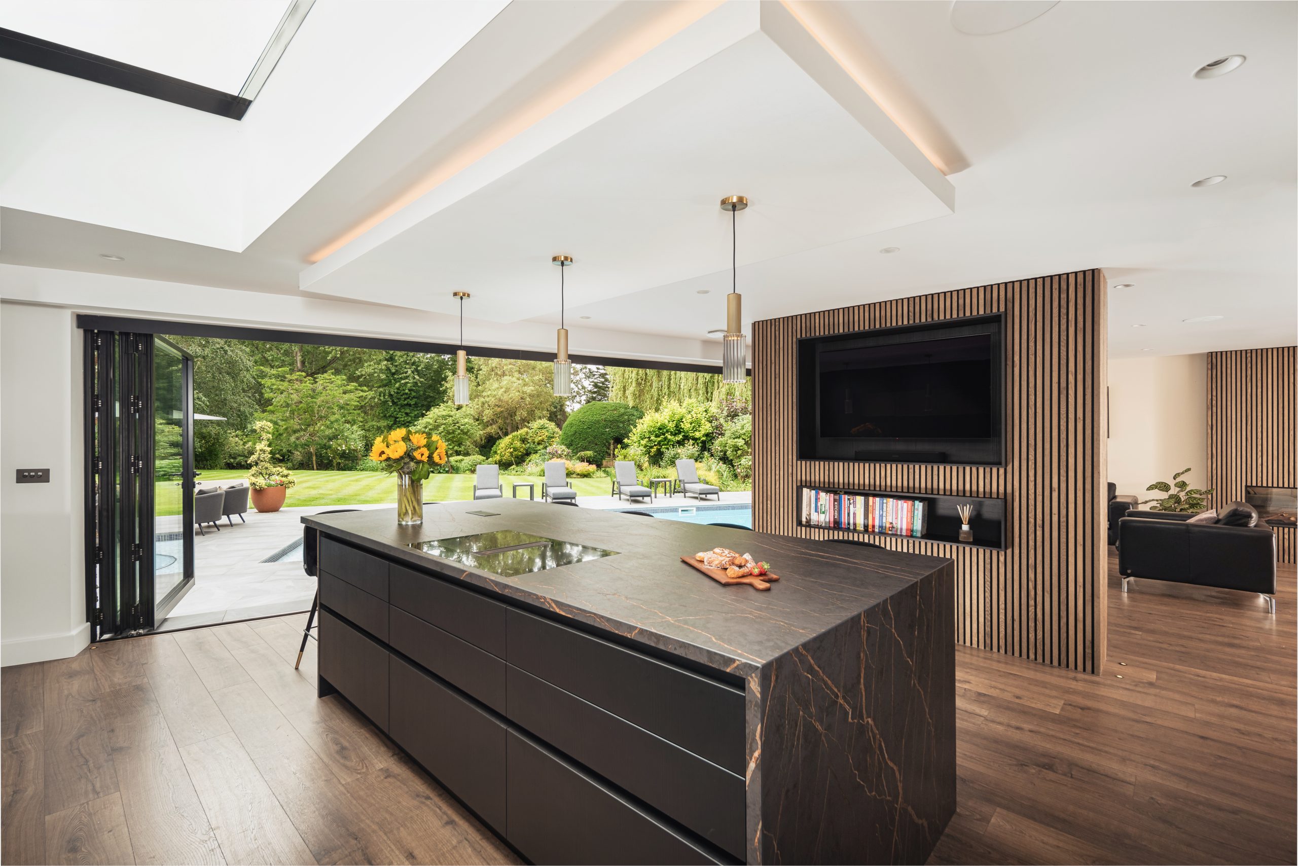 Open-plan kitchen with large island, folding doors to garden, skylight, and wood-panelled TV wall.