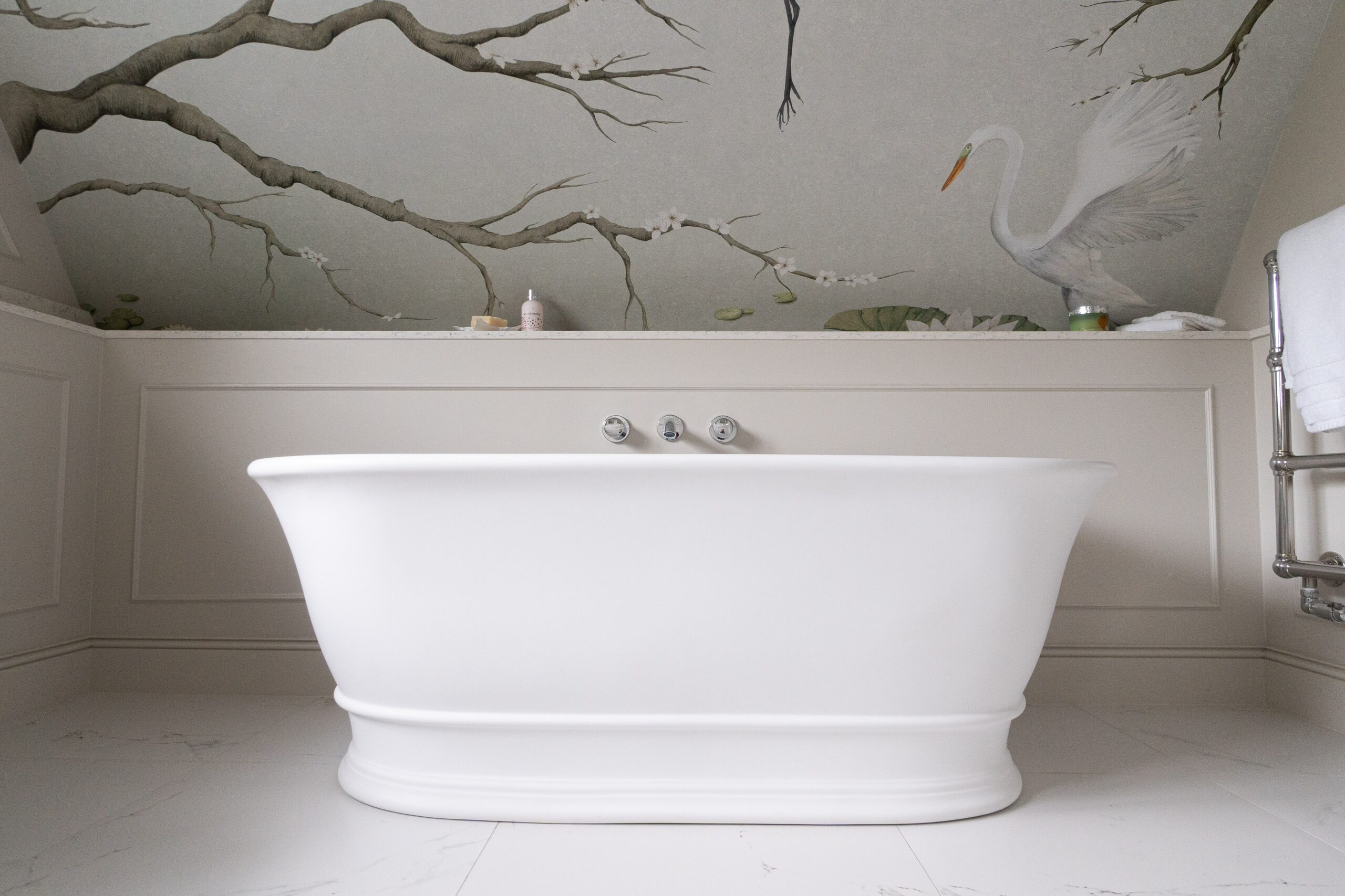 Freestanding white tub with bird and tree mural on the wall, next to a towel rack and marble floor.