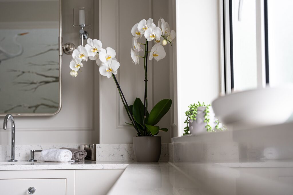 Close-up of white orchids on a marble bathroom counter, with a chrome faucet and rolled towels nearby.