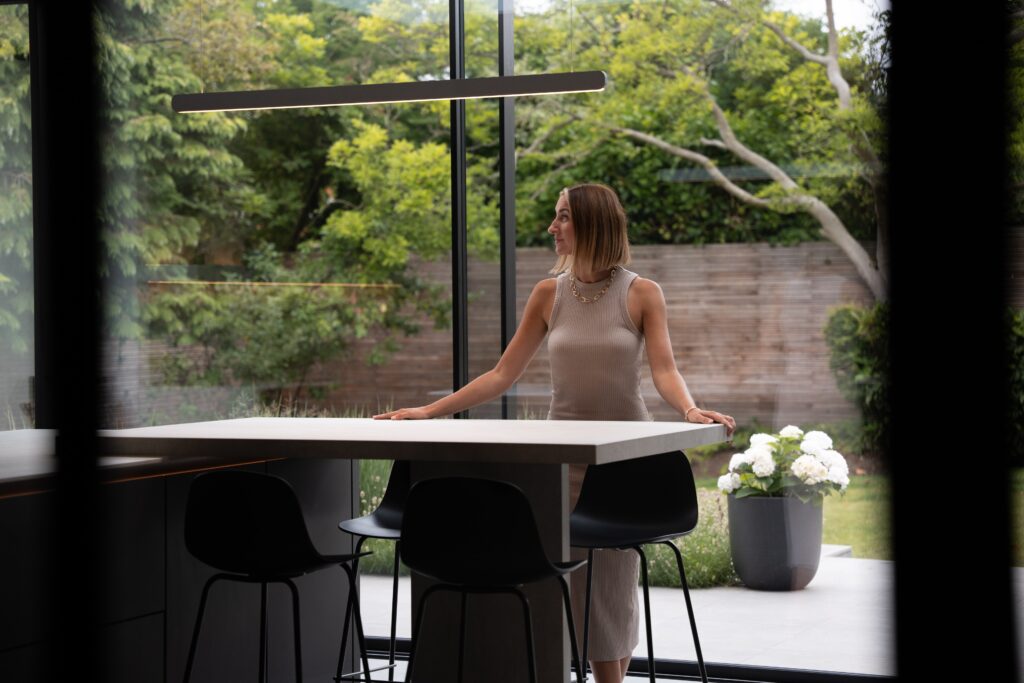 A woman stands by a kitchen island, looking outside through large glass windows to a garden