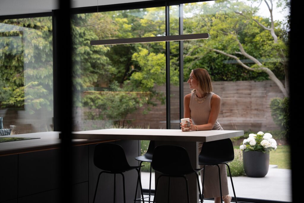 further shot of woman leaning at a modern kitchen island with a mug, back to large glass windows to the garden