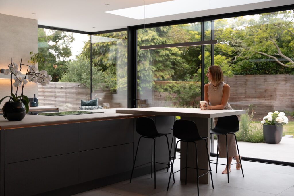 A woman sits at a modern kitchen island with a mug, with back to large glass windows to the garden