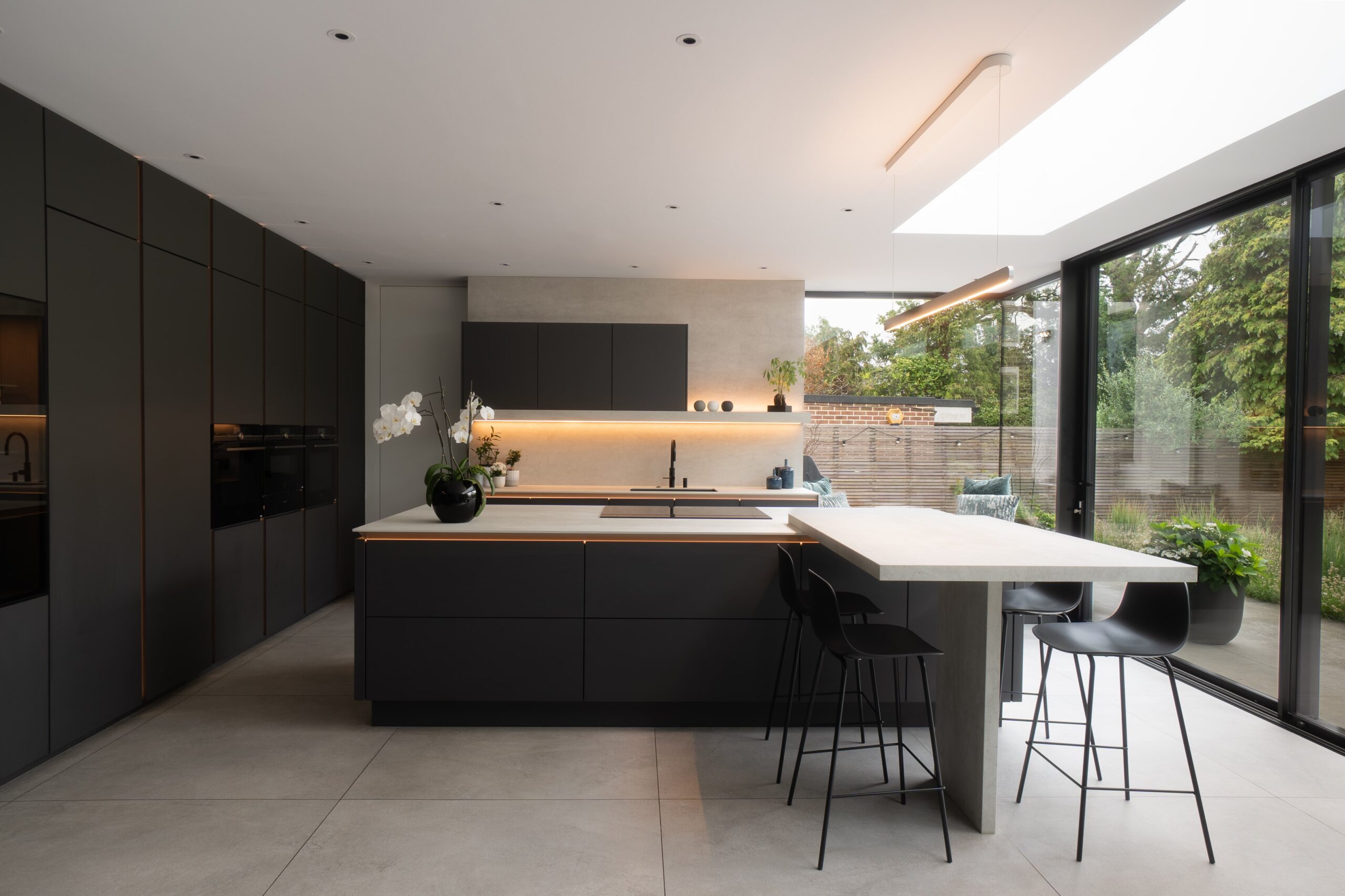 Modern kitchen with dark cabinets, island, bar stools, and large windows overlooking a garden
